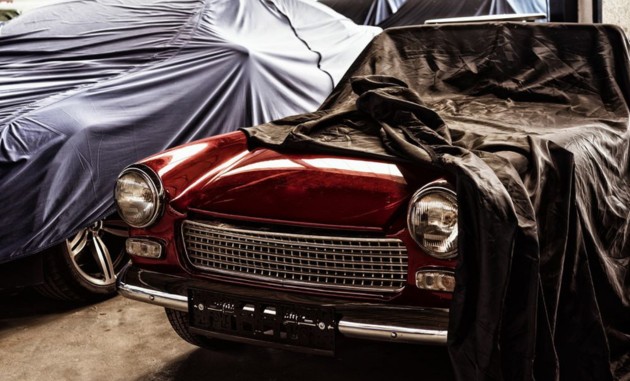 Zwei Oldtimer sind jeweils unter einer Abdeckhaube verborgen, einer ist zur Hälfte aufgedeckt und glänzt in seiner roten Farbe.
